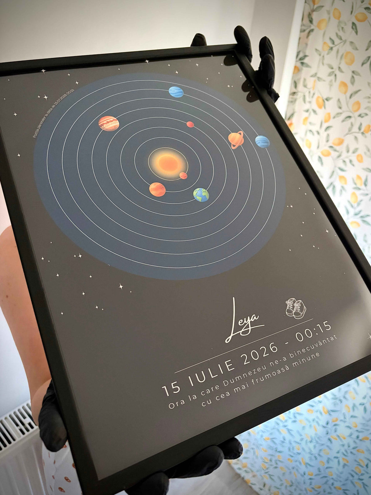 A framed solar system art print held by hands wearing black gloves. The print features a dark gray background with a detailed illustration of the sun and planets in orbit, and personalized text and date printed at the bottom.